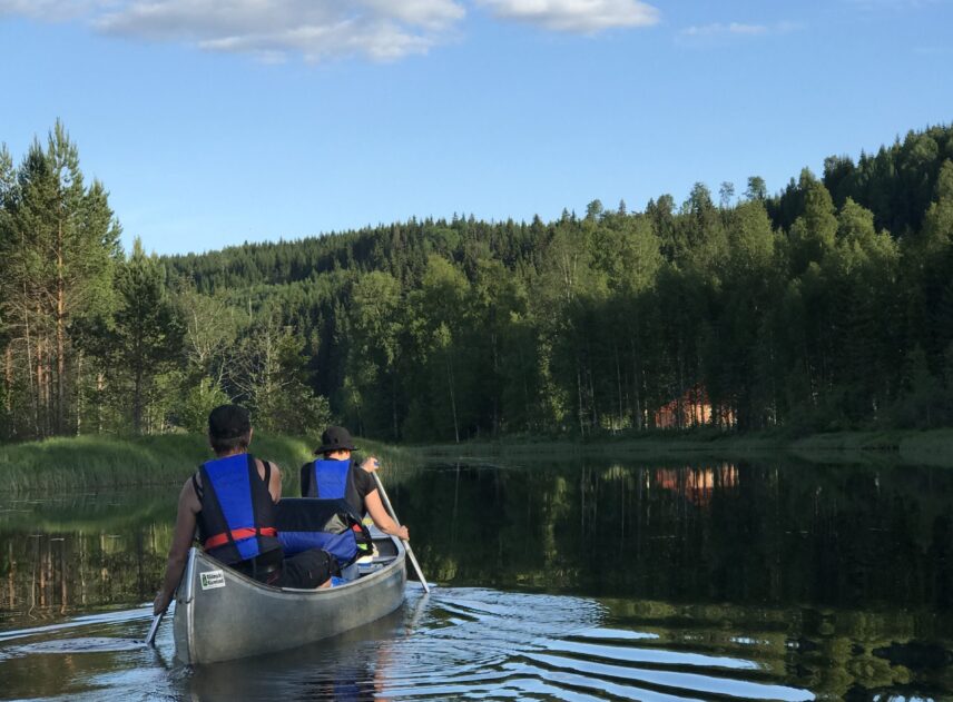 Två personer med flytvästar paddlar kanot. I kanoten ligger också en hel del packning. Skogen är mörk grön, på en ljusblå himmel hänger enstaka vita moln.