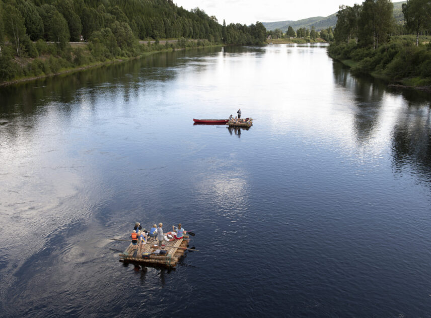En luftbild tagen från hög höjd. Två mindre timmerflotte flyter med lite avstånd till varandra på den breda Klarälven. På flottarna står och sitter flera personer. Älven ligger blått och stilla, en grön skog begränsar vattnet.