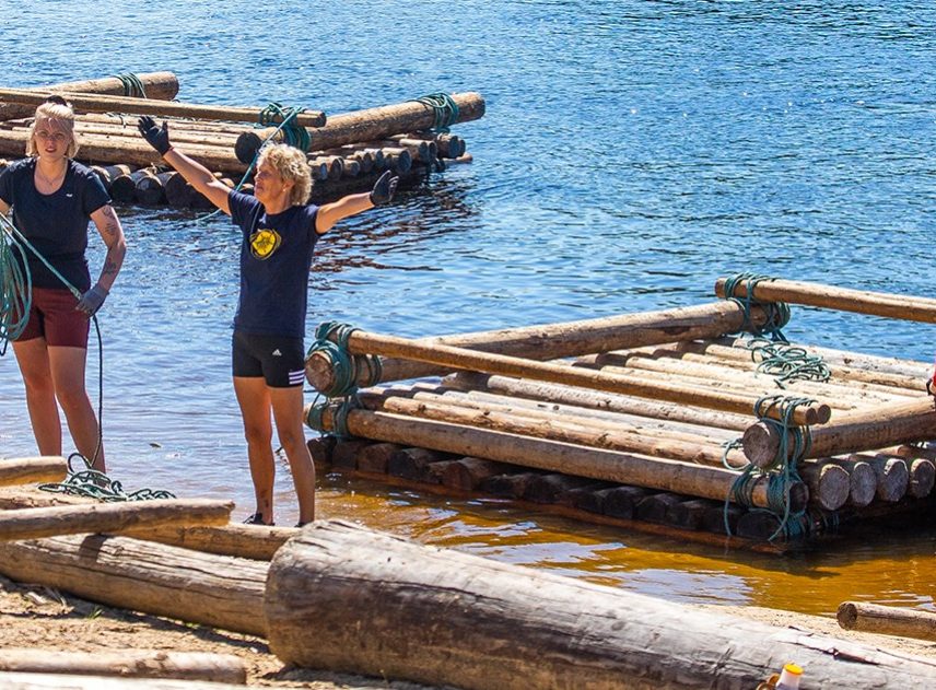 Ett timmerflottbygge i Klarälven har precis avslutats. De färdigbyggda flottarna ligger i vattnet i bakgrunden. En kvinna uttrycker stolt sin glädje över att flotten blev färdig. En annan kvinna håller på att lägga ihop ett rep.
