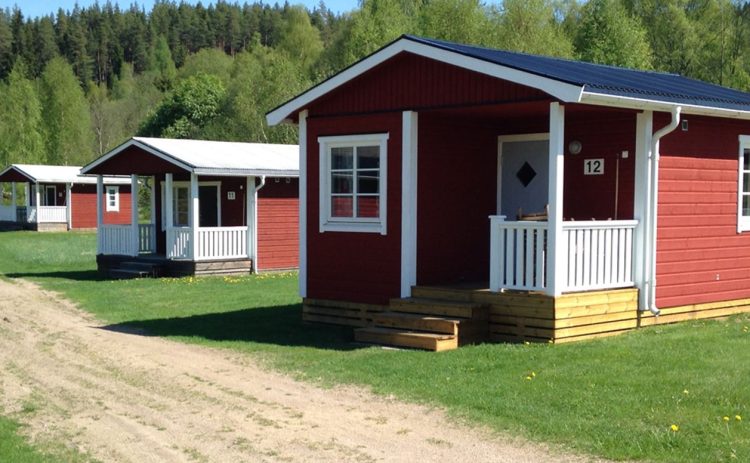 Tre röda stugor med vita verandor ligger i en rad längs en sandväg på Klarälvens Camping. Skuggorna på den gröna gräsmattan visar på en solig dag fast himlen inte är synlig. I bakgrunden breder en grön skog ut sig.