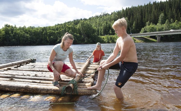 Tre personer håller på att bygger en timmerflotte i Klarälven vid Klarälvens Camping. En pojke tar foten till hjälp för att dra åt repet ordentligt. En kvinna står på knä på flotten och hjälpa till med samma knop. Vid bakre hörnet av flotten står en flicka i vattnet. I bakgrunden leder en bro över älven.
