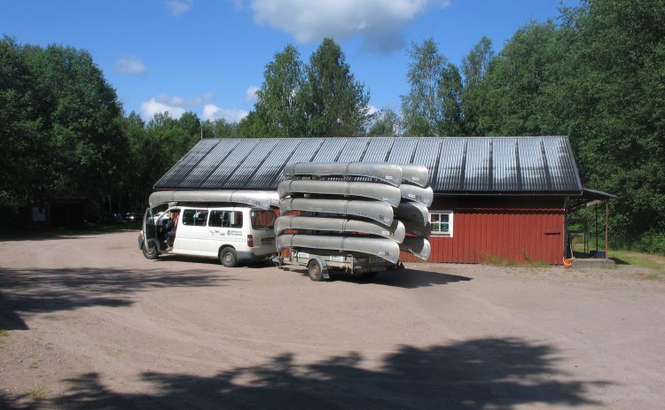 Framför en röd förrådsbyggnad står en vit minibuss med släp. Både bilens tak och trailern är lastade med ett stort antal kanoter. Bilen står i Gunnerud på grusplanen, redo för avfärd.