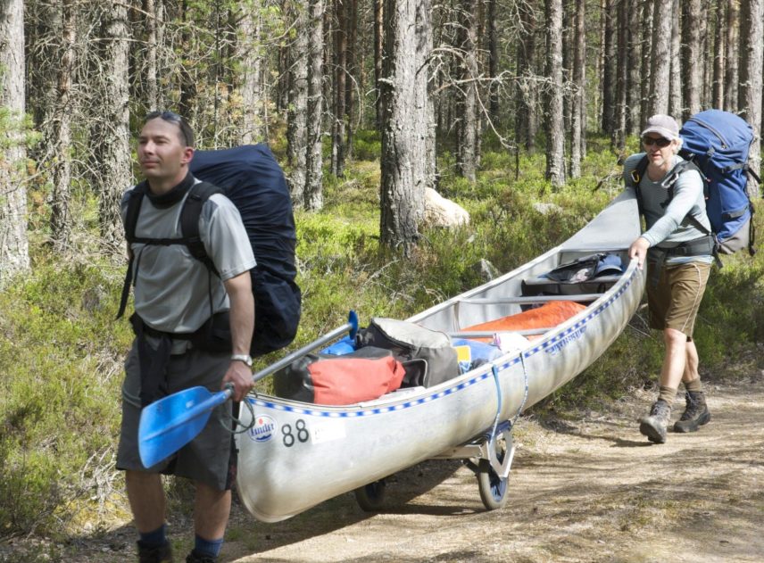 Två personer gör en landtransport av en kanot. Kanoten ligger på en kanotvagn. Personerna bär stora ryggsäckar och och bär eller puttar kanoten längs en sandväg genom skogen.