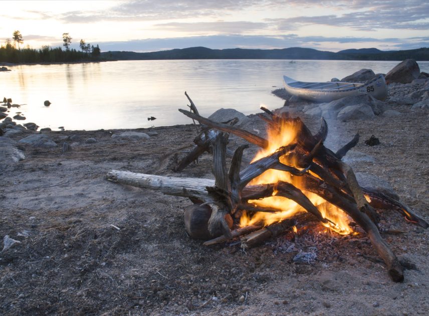 På en sandstrand brinner en brasa. Roten av ett dött träd ligger i lågorna. I vattenbrynet i bakgrunden syns en kanot. Sjön speglar en ljus himmel.