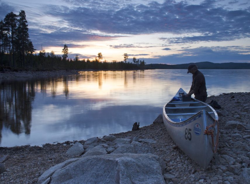 En man står på knä bredvid en kanot som är ligger på en stenig strand. Mannen använder kanotens sits som ett bord medan han diskar. På andra sidan sjön går solen ner, kvällsljuset bryter genom den mörkblå himmel.