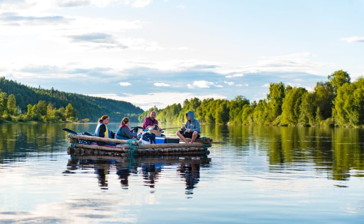 Fyra personer sitter på en timmerflotte utan tältkonstruktion. Klarälven speglar deras kroppar som svarta skuggor på vattnet. Människorna sitter i var sitt hörn på flotten, packningen i mitten. Skogsbeklädda berg i bakgrunden.