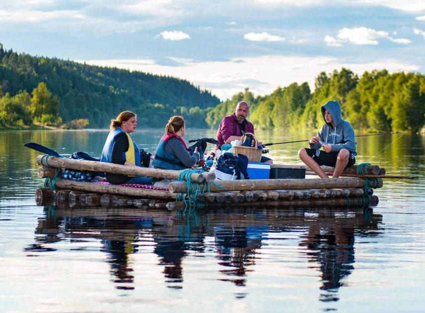 Fyra personer sitter på en timmerflotte utan tältkonstruktion. Klarälven speglar deras kroppar som svarta skuggor på vattnet. Människorna sitter i var sitt hörn på flotten, packningen i mitten. Skogsbeklädda berg i bakgrunden.