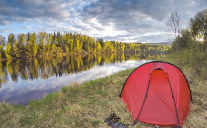 Ett rött Hillebergtält står uppsatt på strandkanten. Utanför ingången i gräset två par skor. Skogen på andra sidan älven speglas tydligt och vattnet visar himlens blågrå molnformation.
