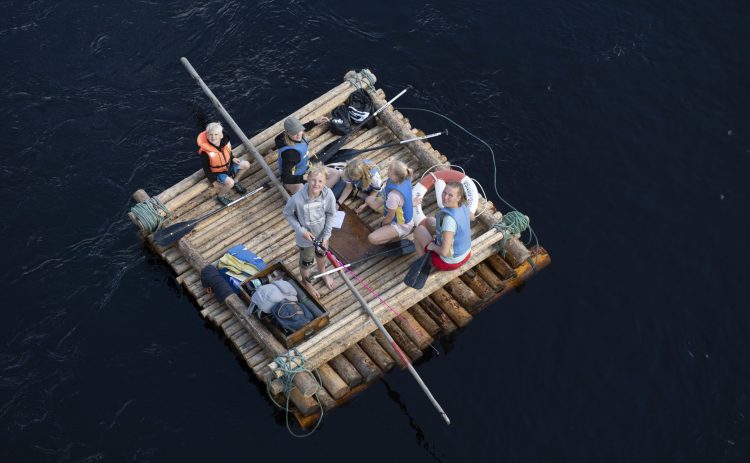 Luftbild på en mindre timmerflotte med flera personer på, både vuxna och barn. Förutom flotten syns bara Klarälvens vatten. Människorna tittar uppåt i kameran, en pojke fiskar från flotten