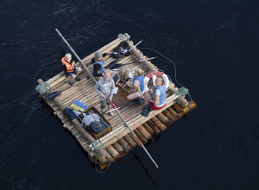 Luftbild på en mindre timmerflotte med flera personer på, både vuxna och barn. Förutom flotten syns bara Klarälvens vatten. Människorna tittar uppåt i kameran, en pojke fiskar från flotten