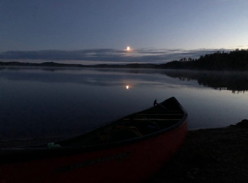 Ovanför en mörk och still sjö lyser månen ikapp sin spegelbild i vattnet. I förgrund syns de svarta konturerna av en kanot med paddel. En smal remsa land skiljer himlen från sjön.
