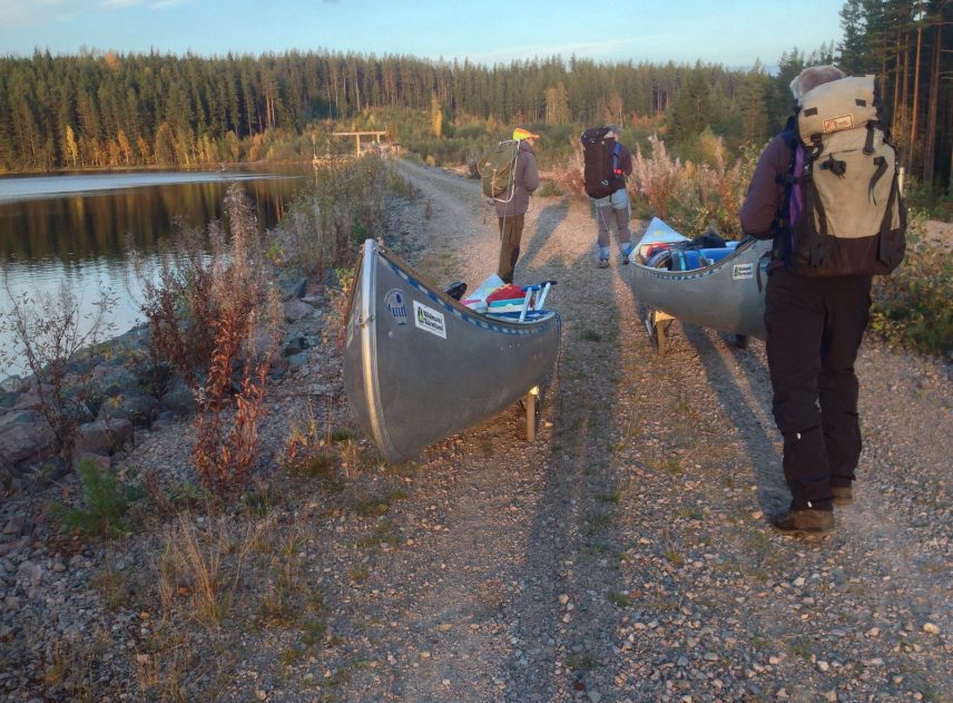 Tre personer går med kanoter på en stenig grusväg längs ett vattendrag. Kanoterna är spända på kanotvagner. Människorna bär stora ryggsäckar och drar eller puttar kanoterna framåt.