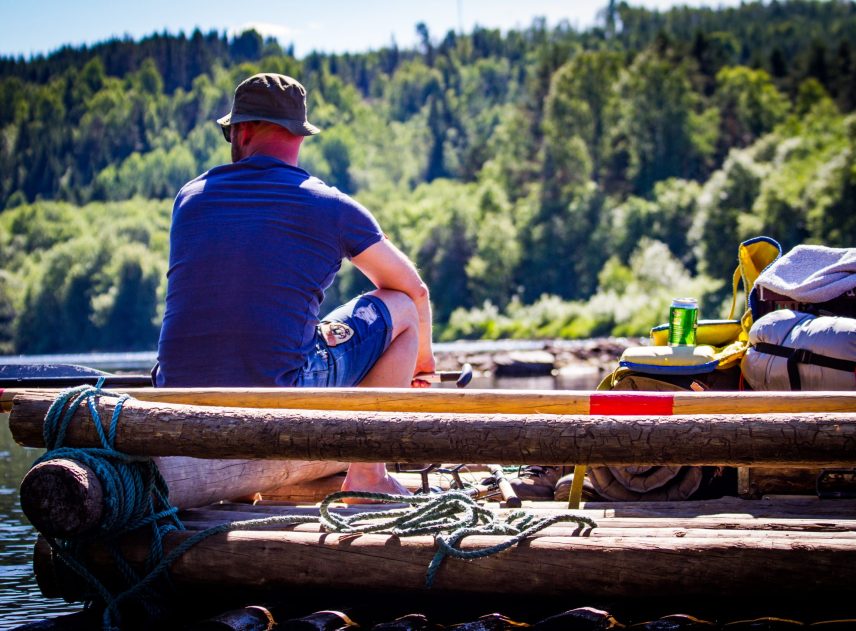 En man med hatt sitter på relingen på en timmerflotte och tittar ner i vattnet. Packningen fyller flottens yta. Flotten flyter på Klarälven, i bakgrunden syns mycket skog.