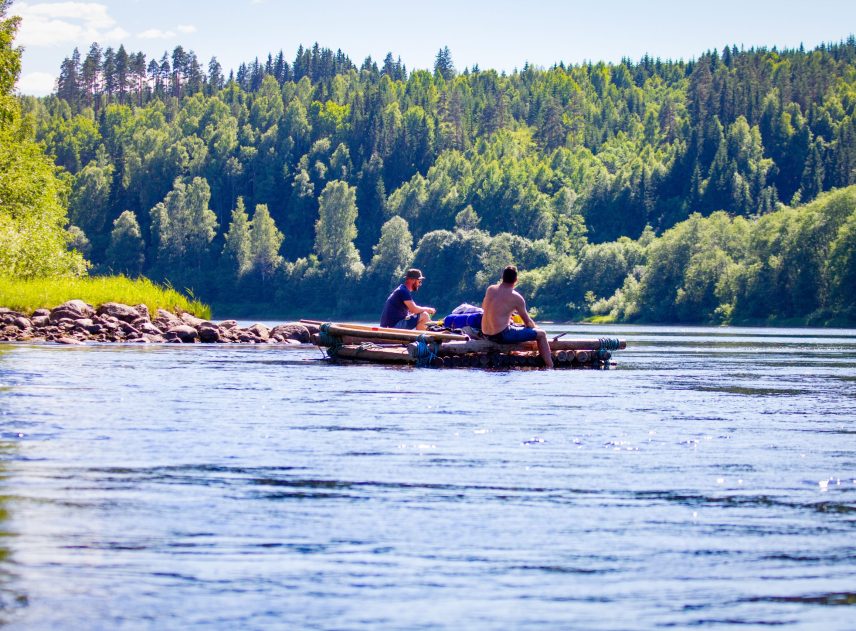 Två män sitter ombord på en mindre timmerflotte utan tältkonstruktion. En varm solig sommardag. Flotte glider lugnt och fint nedströms, männen doppar fötterna i vattnet. Mycket grönska längs strandkanten.