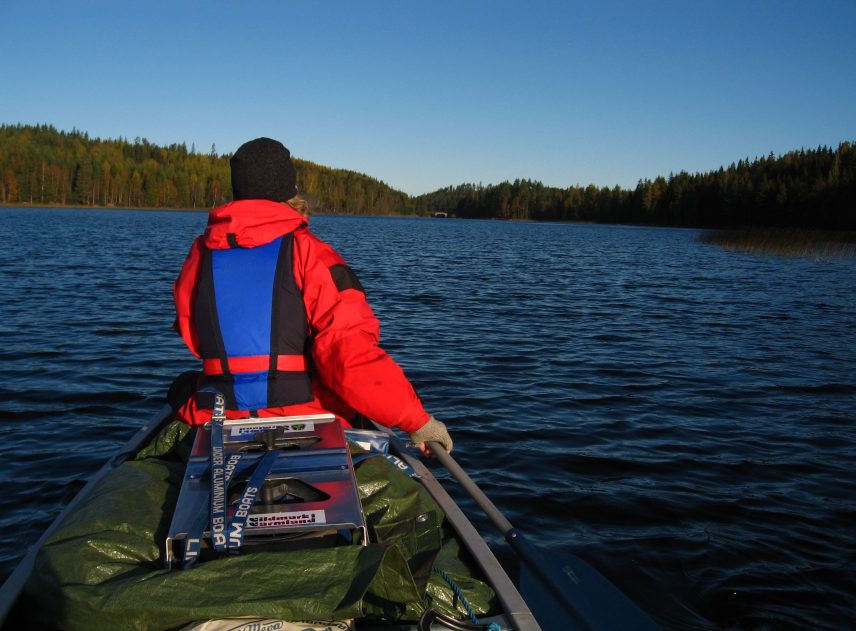 En stor sjö ligger med djupblå vatten. Kanoten syns knappt under packningen som är täckt med en presenning. Däremot är personen med den klarröda jackan väldigt iögonfallande. Sjön begränsas av en tät skog.