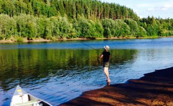 Vid Klarälven står en pojke i vattenbrynet och fiskar. Det är en solig och ljus dag. En kanot som ligger på sanden petar sin spets in bilden. Den andra älvsidan är fullt bevuxen av gröna träd.