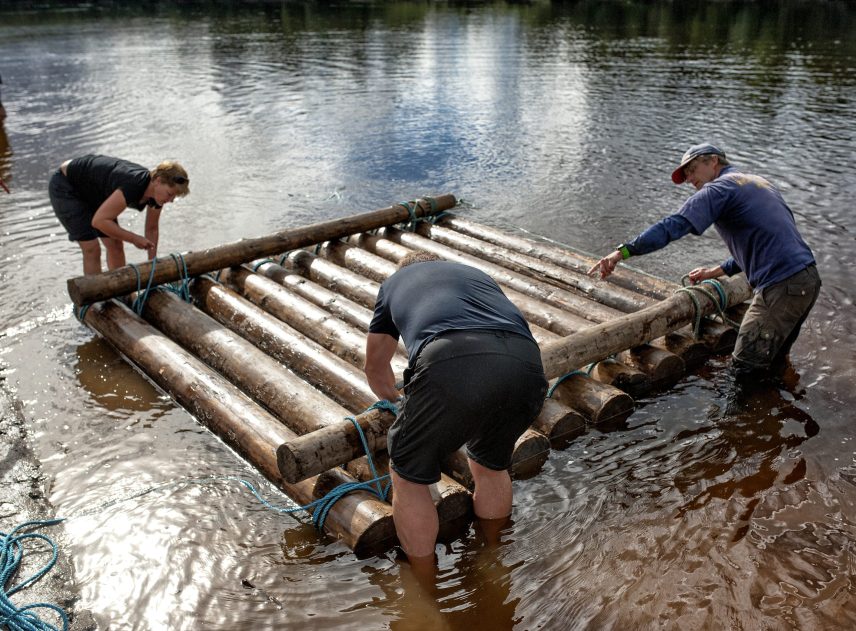 Ett timmerflottsbygge pågår i Klarälven. Tre personer står runt flotten vars första laget redan är färdigbyggd. En man visar de andra två hur de ska göra. Ingen strandlinje är synlig på bilden.