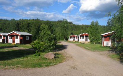 Röda stugor med vita verandaräcken står utspridda på Klarälvens Camping. En grusväg leder till stugorna. En blå himmel är delvis täkt av moln. I bakgrunden syns en grönmättat skog.