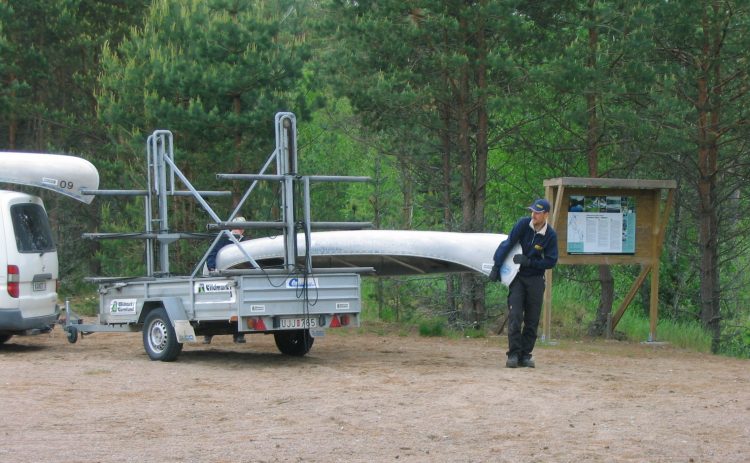 En bil med kanottrailer står på ett grusplan. Två personer håller på att lasta av den sista kanoten från trailern. På bilens tak ligger en kanot kvar. I bakgrunden mycket träd och en informationstavla.
