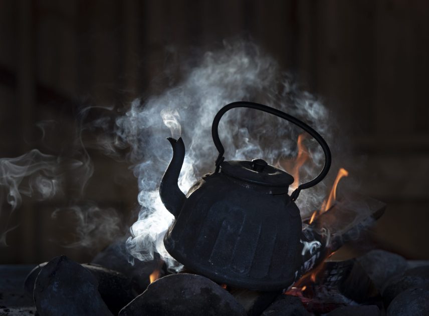 En gammaldags vattenkittel står i en brasa. Konturerna syns tydligt med eldens rök i bakom kaffekannan. Förutom vid lågorna är det en mycket mörk bild, eldstadens stenbegränsning skyms.