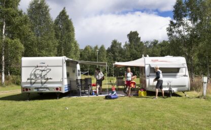 En husbil och en husvagn står mitt emot varann på Klarälvens Camping, förtälten bildar nästan ett genomgående tak. Mellan fordonen syns ett antal människor som tydligen gör sig redo för en flottfärd. Ett barn lekar i gräset, en kvinna bär en flytväst över armen.