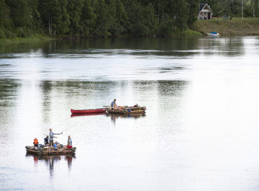 Två mindre flottar utan tältkonstruktion flyter med lite avstånd till varandra på Klarälven. Vattnet blänkar vitt av solen. På ena flotten sitter och står flera personer, en pojke försöker fiska. Andra flotten har med en kanot som jolle. I bakgrunden står ett hus nära älven.