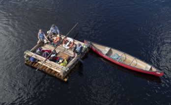 En luftbild av en mindre timmerflotte som flyter på det mörkblå vatten. Förtöjt vid flotten är en röd kanot som är längre än flotten. I kanoten ligger några rep.