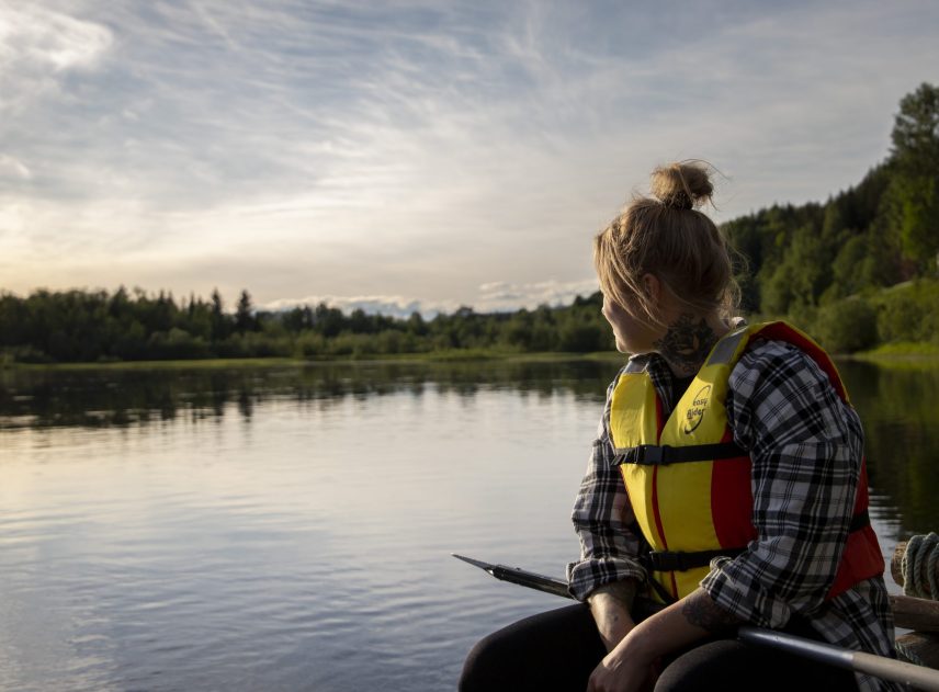En kvinna i flytväst sitter på flotten. Hon är halvt bortvänd och tittar ut över Klarälven. Tvärs över knän ligger en paddel som hon inte behöver just nu. Vattnet är stilla och spegla skogen på motsatt sida älven.