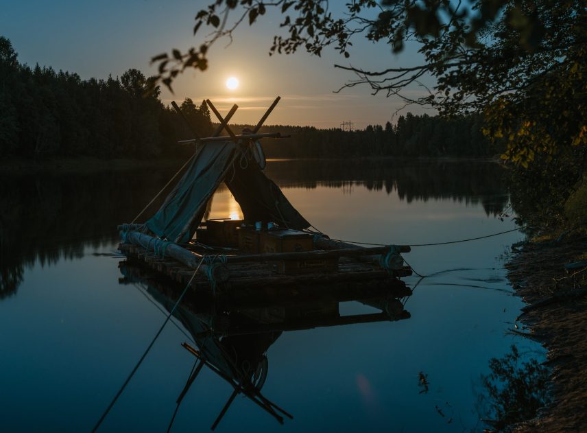 En förtöjt flotte ligger i Klarälven. Himlen och den motsatta strandlinjen är svarta. Solen är på väg ner och lyser genom flottens tältkonstruktion. Bilden ramas in av löven från ett träd.