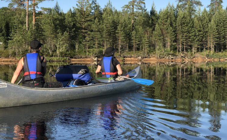 Två personer i svartblå flytvästar paddlar med en fullastad kanot. Det är en varm sommardag. Skogen i bakgrunden är frodig grön.