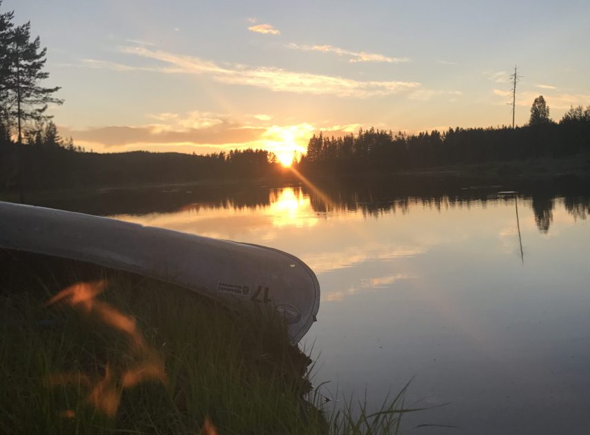 Kvällen har lagt sig över sjön. Snart försvinner solen bakom träden. I förgrund brinner en brasa på stranden. Bredvid ligger en upp och ner vänd kanot. En gråblå himmel speglas i det blanka vattnet.