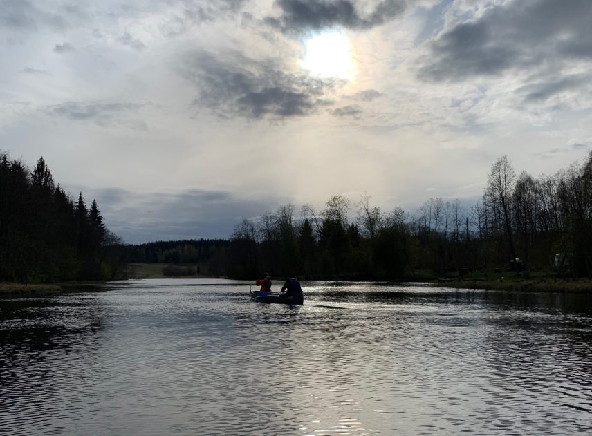 Solen har brutit sig genom det grå molntäcket på himlen. På en skimrande blank sjö syns konturerna av en kanot med två personer i. Vattnet begränsas av en svart skog.