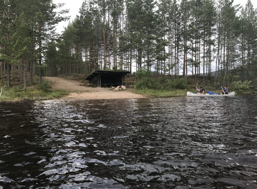En kanot anländer precis vid ett vindskydd vid Skallbergssjön som är slutstation för paddlingen på Rottnan. En bred grusväg leder ända fram till vattnet. Träden står glest på näset.