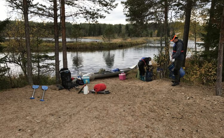 Två personer förbereder paddlingen på Svartälven. Kanoterna ligger redan i vattnet vid stranden i Tyfors, packningen ligger fortfarande utspridd på sanden. Det silvriga vatten delas av en smal remsa med gräs och små buskar.