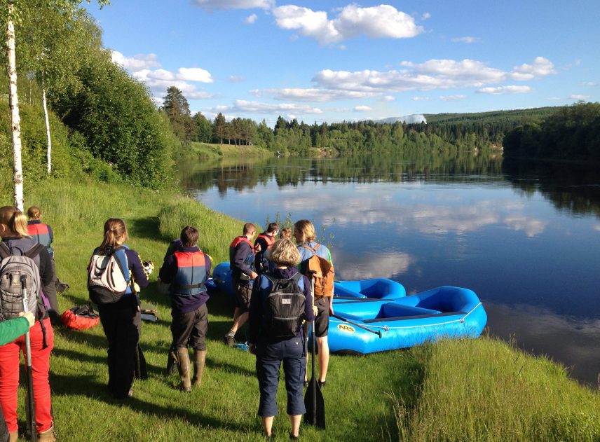 En grupp människor står på gräsmattan vid Klarälven. Redo att skjuva in i vattnet ligger två blå gummibåter. Björkarna står ljusgröna, i älven speglas små lätta moln.