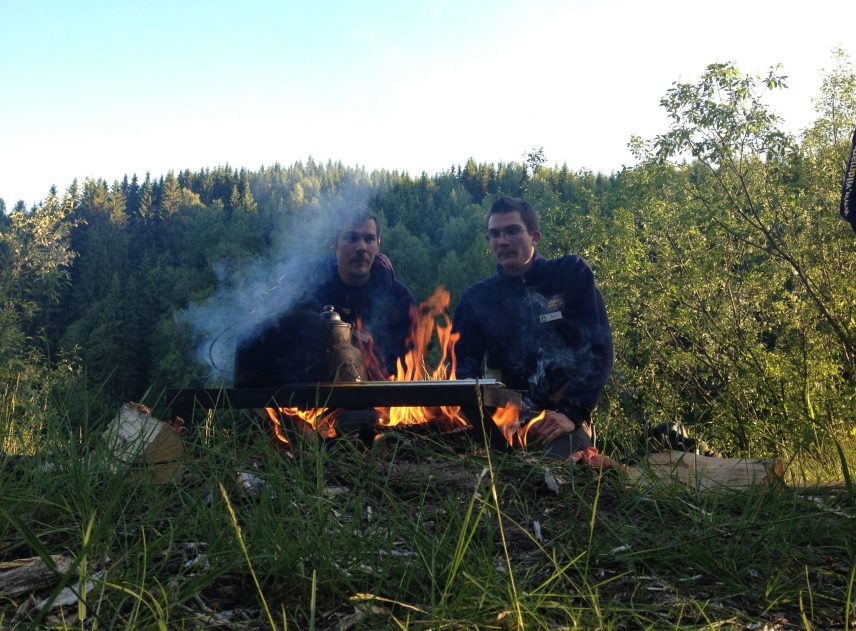 Bakom en brasa sitter två män och tittar in i lågorna. Eldstaden är omgiven av mycket gräs, himlen i bakgrunden lyser ljusblått. På ett grillgaller över glöden står en kaffekittel.