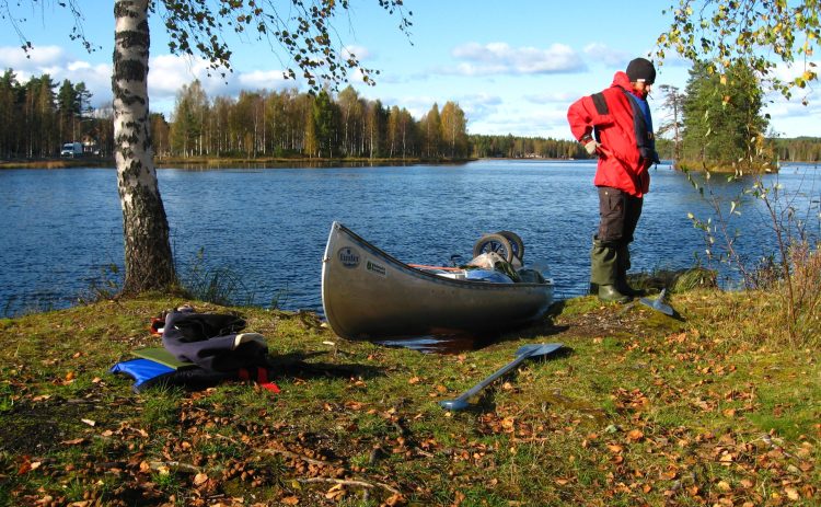 En kanot ligger på en gräsmatta som bildar stranden till en sjö. Höger om kanot står en person i röd jacka, vänster om kanoten står en björk med vit stam. Ljuset verkar komma från en lågt stående sol, första höstlöv på marken.