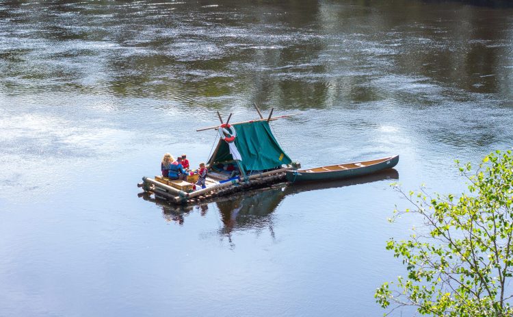 En timmerflotte flytter på Klarälvens glittrande vatten. Stranden syns bara genom speglingen. På flotten sitter två personer och vilar. En kanot flyter fastbunden bredvid flotten.