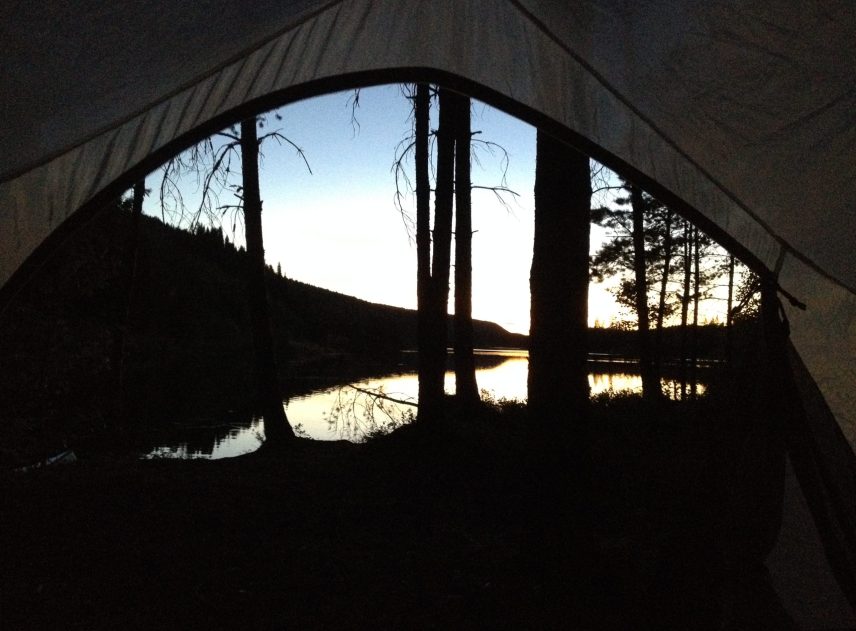 Blicken går genom en tältöppning ut på ett vattendrags som skyms bakom ett antal träd. Kvällsljuset ligger över skogen men himlen är fortfarande blå. En underbar lägerplats i norra Värmland.