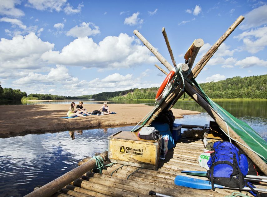 En fullpackad timmerflotte på Klarälven nära en sandbank. På sandbanken sitter tre kvinnor för fika. En strålande sommardag med moln på himlen.
