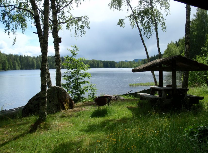 På bilden syns en rastplats vid Röjdån. Ett bord med tak står mitt på en kraftigt grönskande äng. Sjön ligger blankt under en molnig himmel men solen är stark nog för att kasta skuggor över marken.