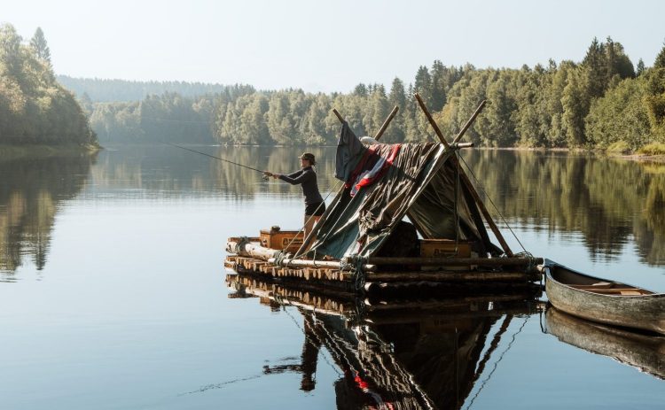 En kvinna står ombord på en flotte i Klarälven och fiskar. Över flottens tältkonstruktion hänger handdukar och annat på tork. Älvens vatten ligger gråblått och spegelblankt. Horisonten bildas av de skogsklädda strandlinjerna.