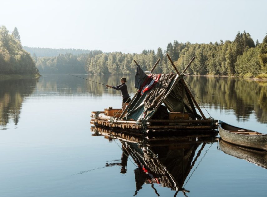 En kvinna står ombord på en flotte i Klarälven och fiskar. Över flottens tältkonstruktion hänger handdukar och annat på tork. Älvens vatten ligger gråblått och spegelblankt. Horisonten bildas av de skogsklädda strandlinjerna.
