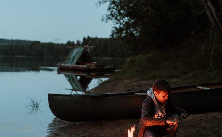 En pojke sitter vid en brasa som är tänd på Klarälvens strand. Bakom honom ligger en kanot uppdragen på land och längre in i bakgrunden syns en timmerflotte med tältkonstruktion. Det är kväll och himlen är på väg att bli mörk.