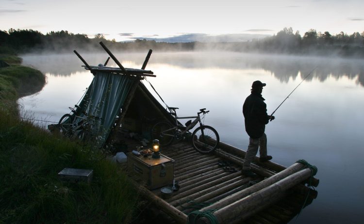 En man står ombord på en stor timmerflotte och fiskar. Klarälvens vatten och strandlinjen ligger i kvällsdimman. På flotten står en ensam liten lampa på en trälåda. Två cyklar syns vid flottens tältkonstruktion.