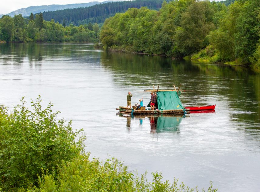 En timmerflotte på Klarälven, bilden är tagen från långt håll. På flotten syns flera personer i färgglada kläder, en person verkar fiska i älven. I bakgrunden skyms bergkedjan. Mycket växtlighet och grönska längs älvkanten, en ljus dag.