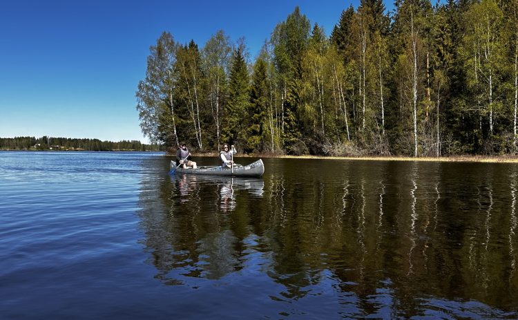Två personer paddlar en kanot på ett stilla vattendrag. Bakom skogen breder en djupblå himmel ut sig. Träden vid stranden speglas i det blå vattnet.