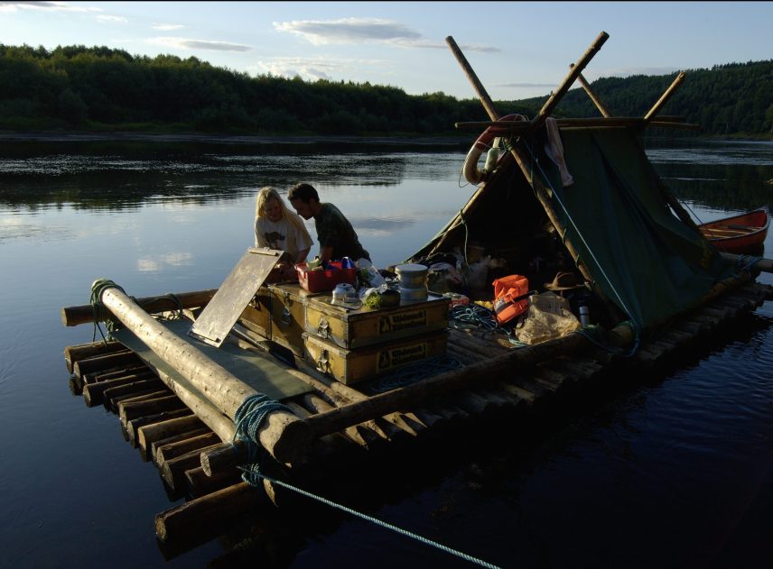 Två personer ombord på en timmerflotte. De verkar leta efter något i en trälåda, kanske förbereder de matlagningen eller disk. Mycket packning under flottens tältkonstruktion. Kan vara en varm sommardag.