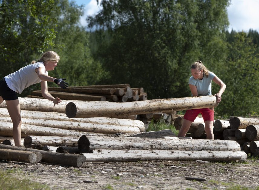 Två kvinnor år på byggplatsen för timmerflottar vid Klarälvens Camping. De samlar ihop stockar för ett flottbygge. Just nu kastar de med mycket kraft en stock som ska användas för flotten.