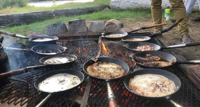 Flera stekpannor ligger på ett stort grillgaller över glöden. Det är en stor eldstad på Klarälvens Camping. I pannorna gräddas smeten för kolbullar. Fläskbitar eller championer ger den extra goda smaken. I bakgrunden syns bron över Klarälven.